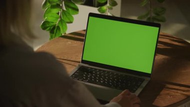 Doğal ışık ve bitkilerle çevrili, ahşap bir masada yeşil ekranlı dizüstü bilgisayarda çalışan bir kadının arka görüntüsü..