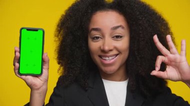 Ceketli mutlu bir kadın elinde yeşil ekranlı bir akıllı telefon tutuyor ve sarı arka plana karşı iyi bir jest yapıyor..
