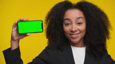 Ceketli, kendine güvenen bir kadın bir elinde yeşil ekranlı akıllı bir telefon tutarken diğer elinde de baş parmağını kaldırıyor..