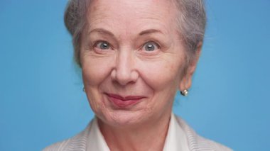 Gri saçlı, inci küpeli, iş elbisesi giymiş mavi bir arka plana içten bir gülümsemeyle bakan son sınıf öğrencisi bir kadının dostane yakın çekimi..