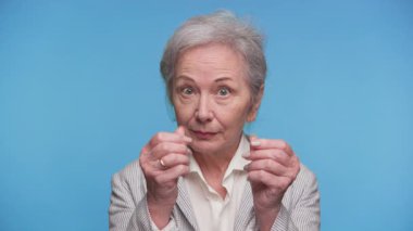 Çizgili ceketli yaşlı bir kadın iki eliyle sağlam bir mavi arka plana karşı para hareketi yapıyor, ciddi görünüyor..