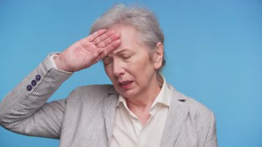 Alnını kapalı tutan çizgili ceketli yaşlı kadın mavi arka planda rahatsızlık ya da ateş belirtileri gösteriyor..