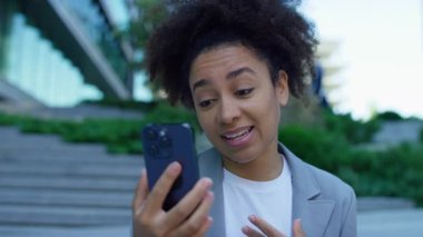 Video görüşmesi sırasında gülümseyen ve konuşan kadın, modern bir şehir ortamında merdivenlerde oturuyor..