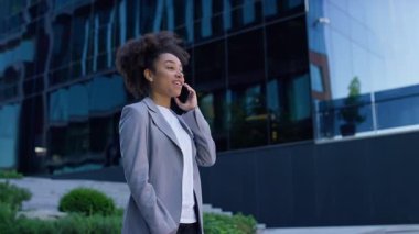 İş kıyafetleri içinde gülümseyen genç girişimci açık havada telefonla konuşuyor, modern cam ofis binasının önünde duruyor..