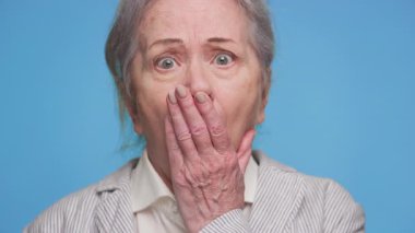 Son sınıf öğrencisi bir kadının eli ağzında, gözleri tamamen kuşkuyla çevrili, mavi arka planda şaşkınlık ya da şok belirtileri gösteren bir fotoğrafı..