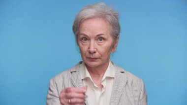 Ceketli yaşlı bir kadın el hareketi yapıyor, para ya da ödeme için işaret veriyor, ciddi bir ifadeyle, mavi arka planda izole edilmiş..