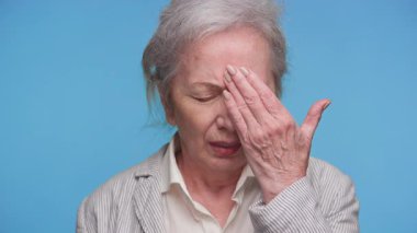 Alnını sıkıntı içinde tutan yaşlı bir kadın, gözleri kapalı, hayal kırıklığını, yorgunluğunu ya da mavi bir arka plana karşı zihinsel yorgunluğunu ifade ediyor..