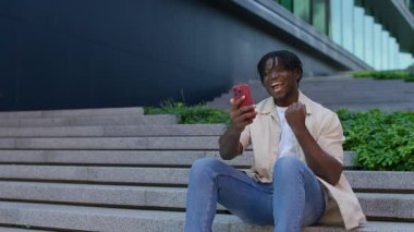 Mutlu genç adam dışarıda merdivenlerde oturuyor, yumruğunu kaldırıyor ve akıllı telefona gülümsüyor başarıyı veya iyi haberi kutlarken..