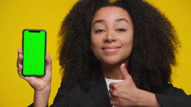 Siyah ceketli gülümseyen kadın yeşil ekranlı bir akıllı telefon tutuyor ve parlak sarı arka plana başparmak işareti veriyor..