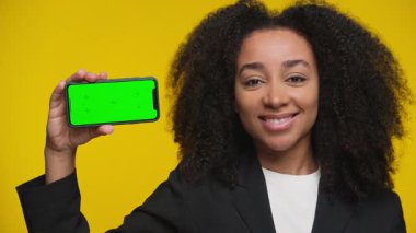 Spor ceketli mutlu kadın yeşil ekranlı akıllı telefonu parlak sarı arka planda tutarken onaylıyor..