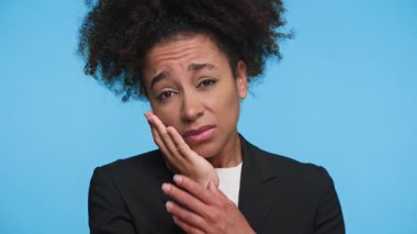 İş elbisesi giymiş genç bir kadın, mavi arka planda diş ağrısı ya da yüz rahatsızlığı olduğunu gösteren acı dolu bir ifadeyle yanağını tutuyor..