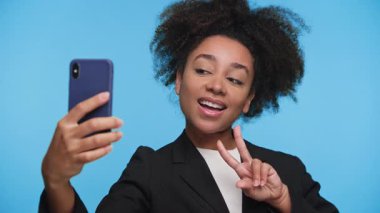 Ceketli gülümseyen genç bir kadın, parlak mavi bir arka planda barış işareti gösterirken akıllı telefonuyla selfie çekiyor..