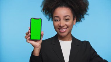 Spor ceketli neşeli bir kadın yeşil ekranlı bir akıllı telefon sunar ve parlak mavi arka plana karşı iyi bir el hareketi yapar..