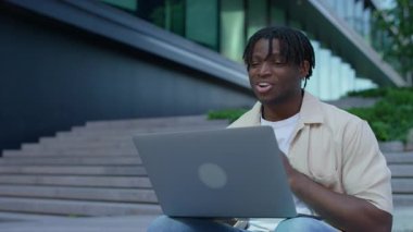 Genç adam dışarıda merdivenlerde dizüstü bilgisayar kullanarak oturuyor, gülümsüyor ve modern şehir ortamında bir video görüşmesi sırasında konuşuyor..