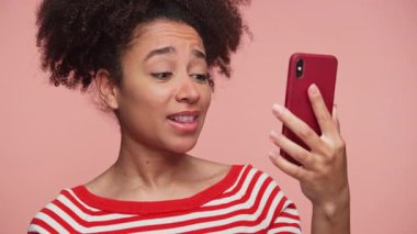 Çizgili tişörtlü neşeli genç bir kadın akıllı telefon görüşmesinde konuşuyor. Telefonunu pembe arka planda tutuyor..