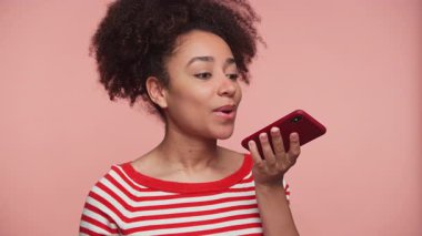 Çizgili tişörtlü genç bir kadın ağzının yanında bir akıllı telefon tutuyor, pembe arka planda bir ses mesajı yolluyor ya da kaydediyor..
