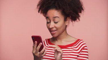 Genç bir kadın akıllı telefonuna bakarken şaşkın görünüyor. Pembe arka planda internet alışverişi yaparken şüphe ve kararsızlığını dile getiriyor..