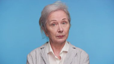 Gri saçlı, çizgili ceketli yaşlı kadın mavi arka plana karşı uyarıda veya ihtiyatlı bir tavırla parmağını kaldırıyor..