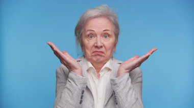 Gri saçlı, çizgili ceketli yaşlı kadın elini kaldırıyor ve mavi arka planda şaşkınlık gösteren, kafası karışık bir yüz ifadesi takınıyor..