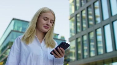 Modern şehir caddesinde yürüyen genç profesyonel kadın, akıllı telefonundaki mesajlara ya da mesajlara odaklanmış..