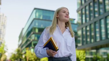 Modern bir iş bölgesinde elinde renkli defterler, çalışmaya ya da çalışmaya hazır genç, profesyonel bir kadın güvenle dışarı çıkıyor..