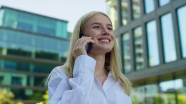 Güneşli bir günde, modern bir iş bölgesinde beyaz gömlekli, gülümseyen genç bir kadın dışarıda telefon görüşmesi yapıyor..