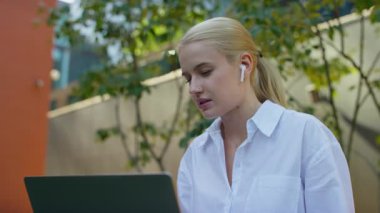 Kablosuz kulaklıklı odaklanmış genç bir kadın, dışarıda modern bir ortamda otururken bilgisayarından yaptığı bir video görüşmesi sırasında konuşuyor..