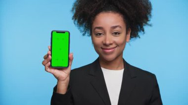 Spor ceketli neşeli bir kadın elinde yeşil ekranlı bir akıllı telefon tutuyor ve başparmak kaldırıyor. Onayı veya olumlu geri bildirimi sembolize ediyor..