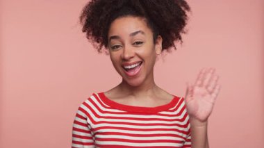 Çizgili tişörtlü mutlu genç bir kadın gülümsüyor ve pembe bir arka plana el sallıyor, arkadaşlığı simgeliyor..