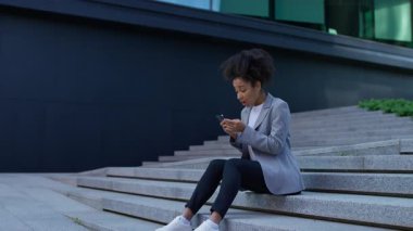 İş kıyafetleri giymiş endişeli bir kadın, dışarıda merdivenlerde oturuyor, telefonda rahatsız edici haberler okurken başını stresle tutuyor..