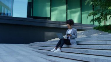 Odaklanmış bir kadın elinde dizüstü bilgisayar ve hesap makinesiyle dış basamaklarda oturuyor, muhasebe, finans veya iş yönetimi görevleri üzerinde çalışıyor.