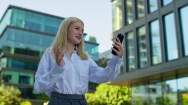 Gülümseyen genç bir kadın akıllı telefondan arıyor. Şehir iş bölgesinde, el sallıyor.