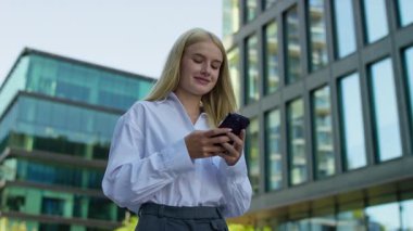 Modern binaların önünde dikilen sıradan genç bir kadın akıllı telefonuyla mesajlaşırken ya da gezinirken hafifçe gülümsüyor..
