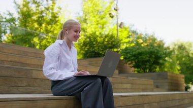 Dışarıdaki tahta basamaklarda dizüstü bilgisayar kullanan, kulaklık takan ve güneşli bir günde yeşil park ortamında çalışan genç bir kadın..
