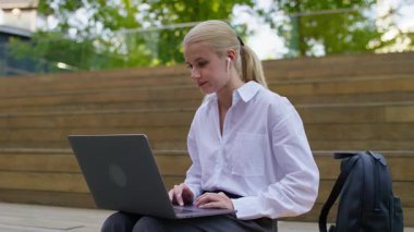 Genç bir kadın dışarıda dizüstü bilgisayarla oturuyor, çalışırken ya da çalışırken yüzünü asık tutuyor, stres ve yorgunluk belirtileri gösteriyor..