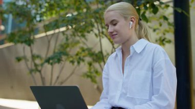 Beyaz gömlek ve kablosuz kulaklık takan genç bir kadın dışarıda dizüstü bilgisayarla oturuyor ve bir video konuşmasında gülümsüyor..