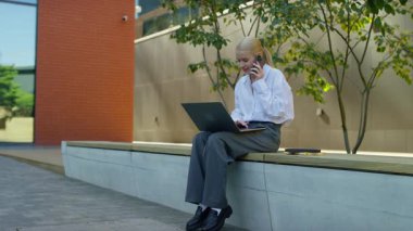 Ofis binasının dışında oturan, dizüstü bilgisayarla çalışırken cep telefonuyla konuşan, görevleri uzaktan yöneten genç profesyonel bir kadın..