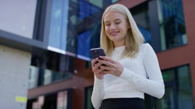 Modern cam binanın yanında duran genç bir kadın güneşli bir günde akıllı telefonuna mesaj yazarken gülümsüyor..