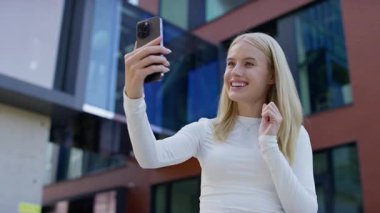 Modern binanın önünde duran beyaz tişörtlü genç bir kadın akıllı telefonuyla video görüşmesi yaparken gülümsüyor ve konuşuyor..