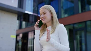 Modern cam binanın yanında duran beyaz tişörtlü genç bir kadın, açık hava telefon görüşmesi sırasında mutlu bir şekilde akıllı telefonuyla konuşuyor..