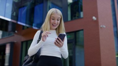 Sırt çantalı genç bir kadın modern binanın önünde duruyor, gülümsüyor ve mutlu bir şekilde akıllı telefonundan olumlu haberler okuyor..