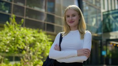 Parlak güneşli bir günde, modern cam binanın dışında güvenle duran, beyaz gömlekli ve sırt çantalı gülümseyen bir kadın..