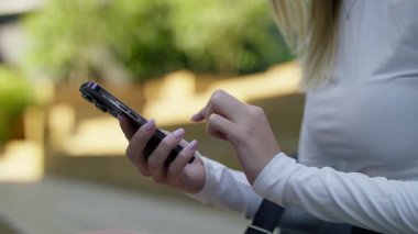 Güneşli bir ortamda arka planda otururken akıllı telefon tutan ve ekranda kayan kadın eller.