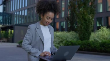 Modern ofis binasının dışında duran kendine güvenen bir iş kadını dizüstü bilgisayarında yazarken gülümsüyor, uzaktan çalışıyor ya da iş yerlerini yönetiyor..