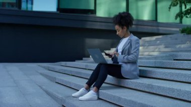 Dış basamaklarda oturan, muhasebe ya da finans işlerini yönetmek için dizüstü bilgisayar ve akıllı telefon üzerinde çalışan odaklanmış genç profesyonel kadın..