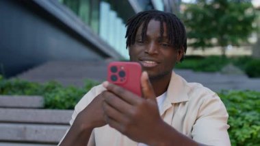 Akıllı telefonunda bir video görüşmesi yaparken gülümseyen genç adam şehir basamaklarının ve yeşilliklerin yanında oturuyor..