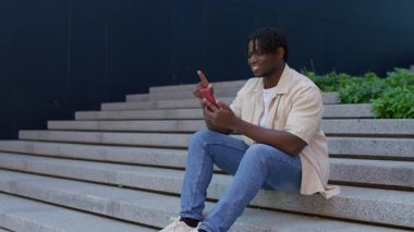 Genç adam dışarıda oturmuş gülümsüyor ve akıllı telefonunu kaydırıyor. Muhtemelen iş ilanları ya da kariyer seçenekleri arıyor..