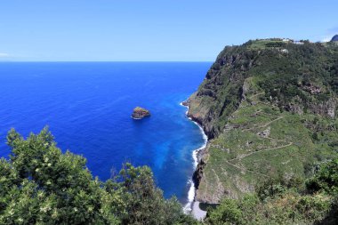 Madeira 'nın kayalık kıyı şeridi. Sarp, sarmal yolu olan yeşil bir kıyı şeridi derin mavi sularla çevrili okyanusa iniyor. Uzakta, denizden yükselen küçük kayalık bir ada var..