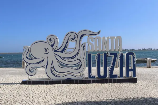 Santa Luzia, Portekiz 'deki su kıyısında asfalt bir gezinti güvertesinde bulunan stilize edilmiş ahtapot çizimi ile SANTA LUZIA' yı gösteren sanatsal beyaz ve mavi bir tabela. Arkaplanda açık mavi gökyüzü.