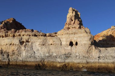 Güney Portekiz 'deki Algarve kıyısı boyunca uzanan kayalık manzaralı bir uçurum, sıcak güneş ışığıyla yıkanmış. Engebeli jeolojik katmanlar doğal gölgelerle vurgulanırken, aşağıdaki sakin turkuaz su açık mavi gökyüzünü yansıtıyor. A